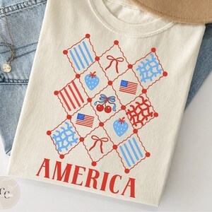 Puede incluir: Camiseta de color crema con un diseño patriótico. El diseño presenta gráficos rojos, blancos y azules, incluyendo fresas, cerezas, lazos y banderas americanas en una cuadrícula. La palabra "AMERICA" está impresa en rojo en la parte inferior.
