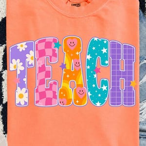 Könnte beinhalten: Ein leuchtend orangefarbenes T-Shirt mit kurzen Ärmeln und dem Wort TEACH in großen, bunten, gemusterten Blockbuchstaben. Jeder Buchstabe hat ein einzigartiges Design mit Gänseblümchen, rosa Karomuster, orangefarbenen Herzen und Sternen.
