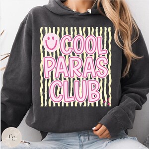 May include: Dark gray hoodie with the text "COOL PARAS CLUB" in pink, outlined in white, and a smiley face. The text is set against a background of black and yellow vertical stripes. The hoodie has a drawstring hood and long sleeves.