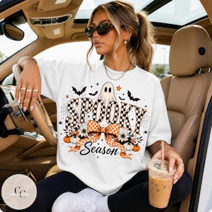May include: A white short-sleeved t-shirt featuring a graphic print with the text SPOOKY SEASON in a black and white checkered pattern. The design includes a ghost, bats, spiderwebs, flowers, pumpkins, and an orange and white checkered bow.