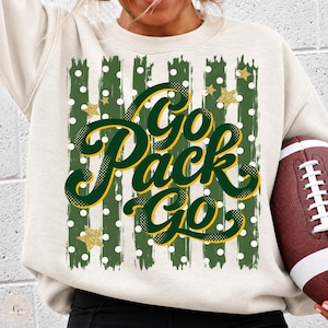 May include: A cream-colored crewneck sweatshirt featuring a graphic print with the text "Go Pack Go" in dark green and yellow script. The design includes vertical dark green brushstroke stripes, white polka dots, and gold glitter stars.
