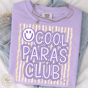 May include: A lilac Comfort Colors crewneck sweatshirt with the text "COOL PARAS CLUB" in a purple font, a smiley face, and gold wavy lines. A gold necklace is draped across the shirt. The shirt is folded on top of a pair of blue jeans.