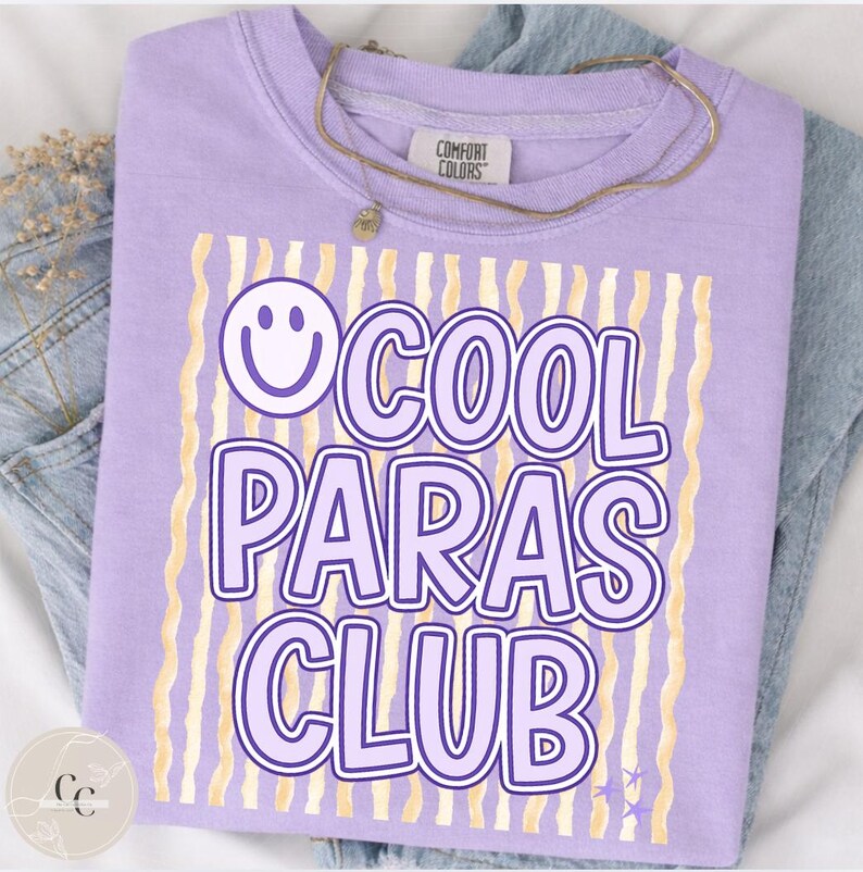 May include: A lilac Comfort Colors crewneck sweatshirt with the text "COOL PARAS CLUB" in a purple font, a smiley face, and gold wavy lines. A gold necklace is draped across the shirt. The shirt is folded on top of a pair of blue jeans.