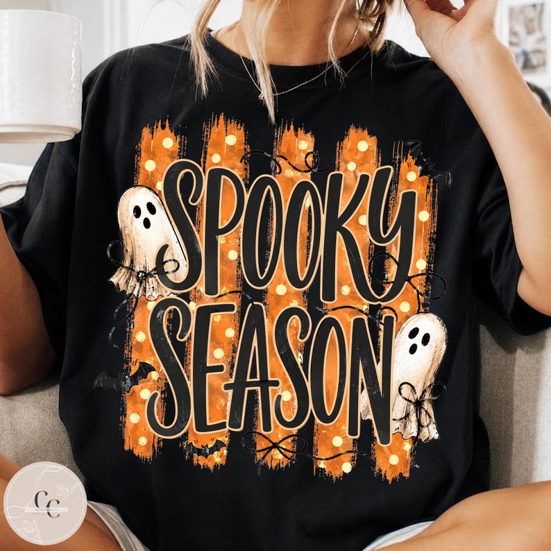 May include: A black t-shirt featuring a graphic print with the text SPOOKY SEASON in large black letters. The design includes vertical orange brushstroke stripes, small white polka dots, two white ghost illustrations, and small black bats.
