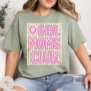 May include: Sage green t-shirt featuring the pink text "GIRL MOMS CLUB" on a background of yellow and white stripes. A pink smiley face is above the word "GIRL".
