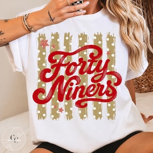 May include: A white short-sleeved t-shirt featuring a graphic design with vertical gold brushstroke stripes and white polka dots. The text Forty Niners is printed in a large, bold red script font, accented by small red glittery stars.