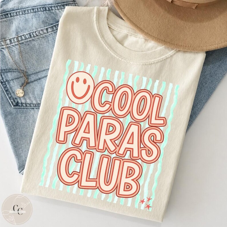 May include: Beige t-shirt with the text "COOL PARAS CLUB" in red and orange, with a smiling face graphic. The design includes light blue vertical stripes and two sets of red stars. The shirt is displayed with blue jeans and a brown hat.