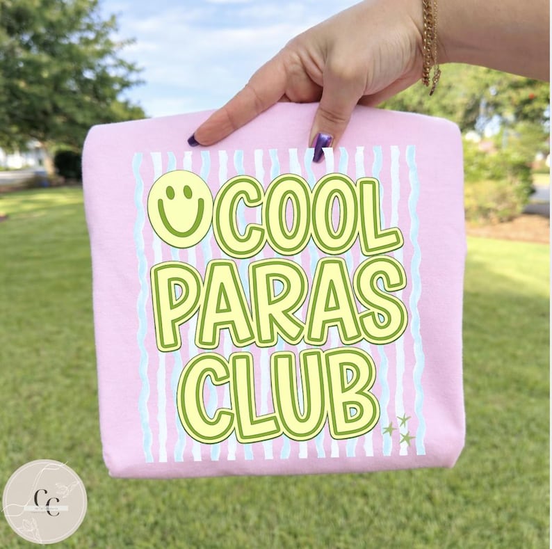 May include: A light pink t-shirt with the text "COOL PARAS CLUB" in a green and yellow font. The design includes a smiley face and vertical stripes in light blue and white. The shirt is held up by a hand.