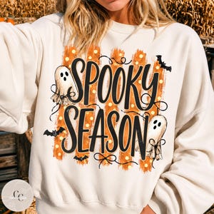 May include: A cream-coloured crewneck sweatshirt featuring a graphic print with the text SPOOKY SEASON in black script. The design includes two white ghosts, three small black bats, and vertical orange brushstroke stripes with yellow dots.