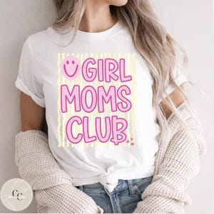 May include: White t-shirt with the text "GIRL MOMS CLUB" in pink letters with a smiley face and stars. The shirt has a light yellow striped background. The model is wearing blue jeans and a cream-coloured sweater.