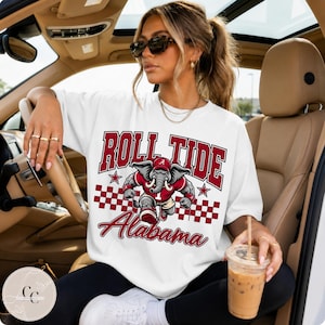 May include: A white short-sleeve t-shirt featuring a graphic print with the text Roll Tide Alabama in maroon lettering. The design includes a cartoon elephant mascot wearing a cap and jersey, surrounded by maroon stars and a checkered pattern.