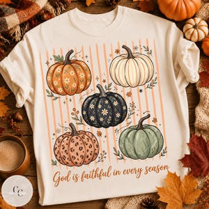 Peut inclure: Un t-shirt crème avec un motif de cinq citrouilles orange, blanches, noires et vertes, ornées de fleurs et d'imprimés léopard. Le fond présente des rayures verticales orange et le texte God is faithful in every season est inscrit en bas.