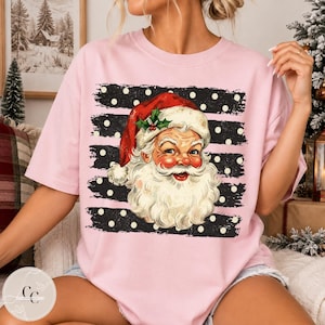 May include: A light pink t-shirt featuring a graphic of Santa Claus wearing a red hat with holly. The graphic is set against a black background with horizontal white stripes and white polka dots, creating a festive holiday design on the shirt.