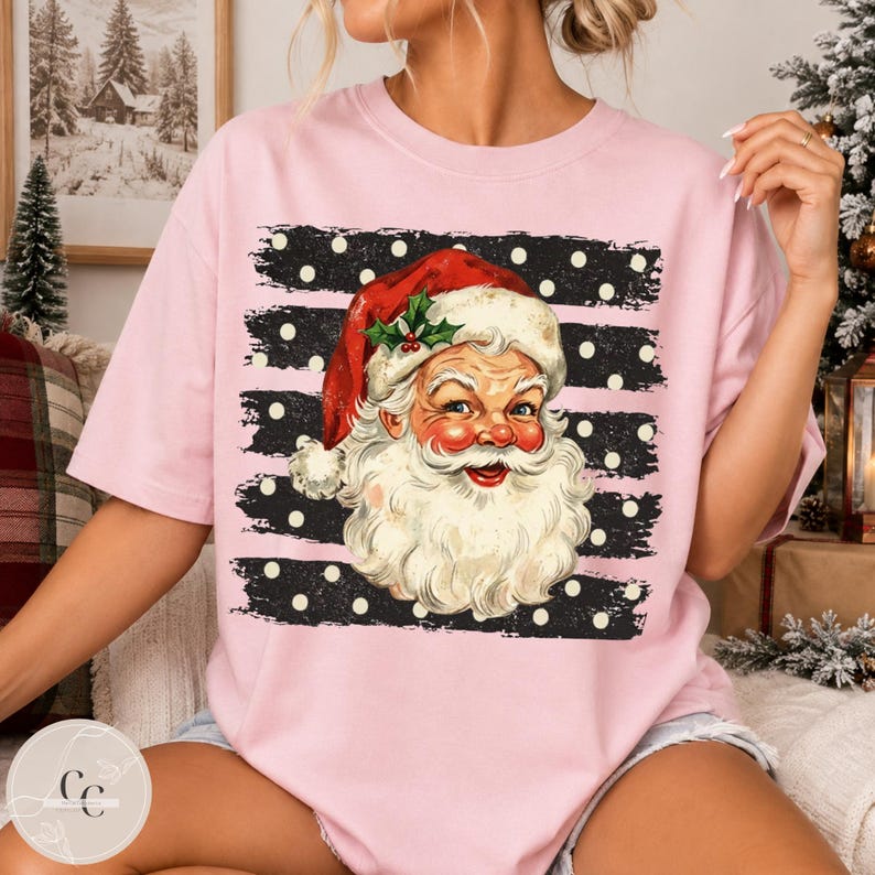 May include: A light pink t-shirt featuring a graphic of Santa Claus wearing a red hat with holly. The graphic is set against a black background with horizontal white stripes and white polka dots, creating a festive holiday design on the shirt.