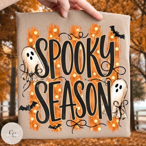 May include: A tan t-shirt featuring the black text SPOOKY SEASON in a stylised font. The design includes orange brushstroke stripes with white polka dots, two white ghosts with black facial features, and three small black flying bats.