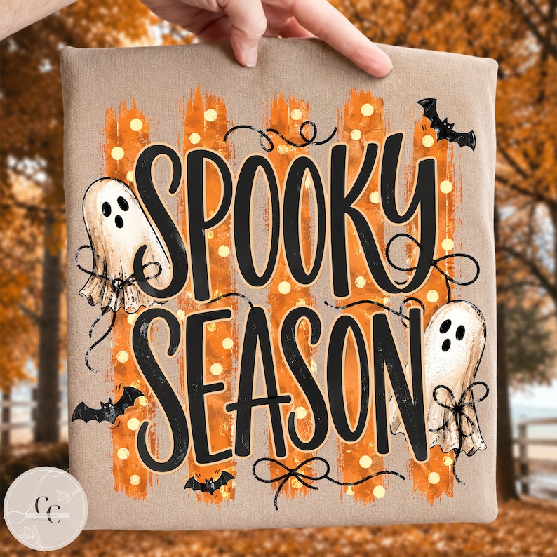 May include: A tan t-shirt featuring the black text SPOOKY SEASON in a stylised font. The design includes orange brushstroke stripes with white polka dots, two white ghosts with black facial features, and three small black flying bats.