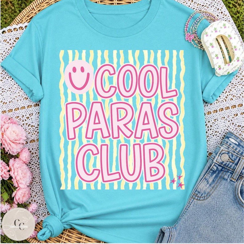 May include: A turquoise t-shirt with the text "COOL PARAS CLUB" in pink letters, a pink smiley face, and yellow and white striped background. The shirt is folded and has a knot at the bottom. Accessories and denim shorts are visible.