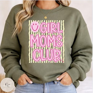 May include: An olive green sweatshirt featuring the pink text "GIRL MOMS CLUB" with a smiling face design. The text is set against a background of vertical yellow stripes. The sweatshirt is a casual, comfortable style.