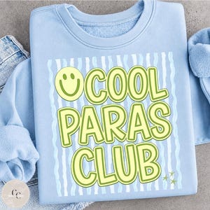 May include: A light blue sweatshirt with the text "COOL PARAS CLUB" in a yellow and green font. The design includes a smiley face and wavy lines. The sweatshirt is made of a soft material.