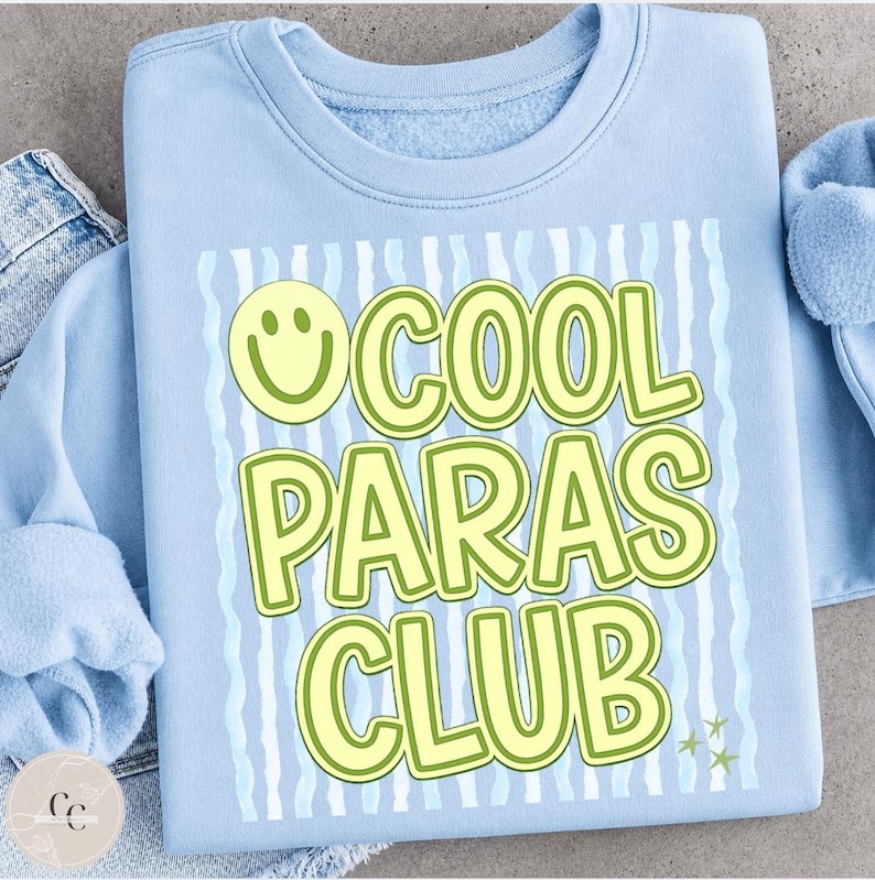 May include: A light blue sweatshirt with the text "COOL PARAS CLUB" in a yellow and green font. The design includes a smiley face and wavy lines. The sweatshirt is made of a soft material.