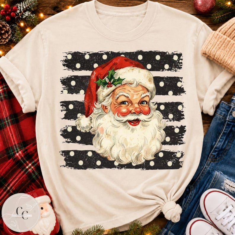 May include: A cream-colored short-sleeve t-shirt featuring a graphic of Santa Claus with a red hat and white beard. The design is set against a black background with horizontal distressed stripes and white polka dots, creating a festive holiday look.