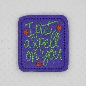 May include: Purple felt patch with green embroidered text that reads "I put a spell on you" and orange embroidered flowers.