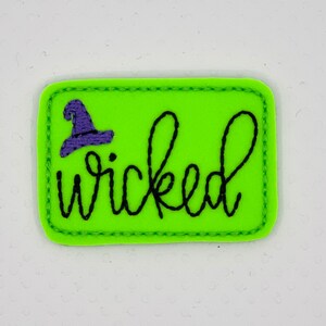 May include: A green felt patch with a black stitched outline and the word "wicked" in black cursive script. A purple witch's hat is stitched above the word.