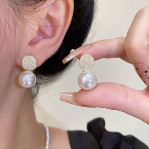 Può includere: Orecchini pendenti con perle. Ogni orecchino presenta una piccola parte superiore ovale ricoperta di cristalli scintillanti, collegata a una grande perla rotonda e lucente. Un accessorio classico per aggiungere un tocco di raffinatezza.