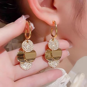 Può includere: Orecchini pendenti color oro con una parte superiore circolare incastonata con piccoli cristalli scintillanti. Sotto, pendono due pendenti circolari piatti color oro, creando un effetto a strati. Gli orecchini hanno una chiusura a cerchio.