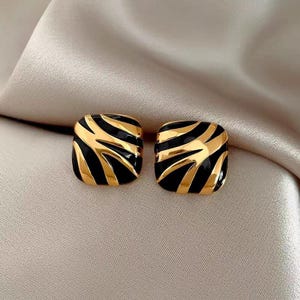 Può includere: Un paio di orecchini quadrati con un design a righe nere e oro. Gli orecchini hanno una finitura lucida e sono presentati su uno sfondo neutro. Un accessorio di moda.