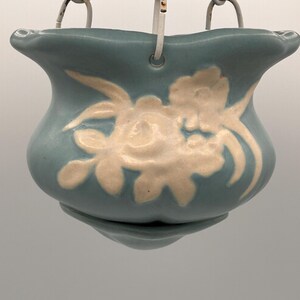 May include: A light blue ceramic hanging planter with a scalloped rim and a raised white floral relief pattern. The planter is suspended by three white metal chains attached to small holes along the upper edge of the bowl-shaped vessel.