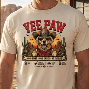 K&ouml;nnte beinhalten: Beiges T-Shirt mit einem Aufdruck, der einen Hund mit Cowboyhut und Sonnenbrille zeigt, dem Text "Yee Paw" und dem Satz "Good Times Cold Drinks No Bad Days". Das Shirt hat auch Symbole f&uuml;r BBQ's, Country-Konzerte, Ausfl&uuml;ge zum See und kaltes Bier.