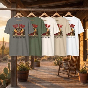 K&ouml;nnte beinhalten: F&uuml;nf kurz&auml;rmelige T-Shirts in verschiedenen Farben: Grau, Olivgr&uuml;n, Off-White, Beige und Wei&szlig;. Jedes Shirt zeigt eine Grafik eines Hundes mit Cowboyhut und dem Text "Yee Paw". Die Shirts h&auml;ngen an Holzkleiderb&uuml;geln.
