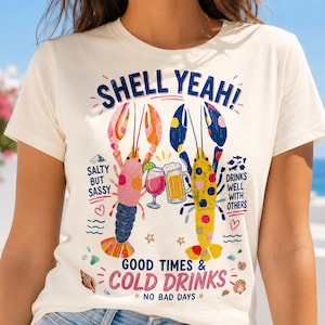 Op de afbeelding: Crèmekleurig T-shirt met een kleurrijk ontwerp van twee kreeften met drankjes. De tekst "SHELL YEAH!" staat bovenaan, met "Salty But Sassy" en "Good Times & Cold Drinks" eronder. Het ontwerp bevat harten, golven en sterren.