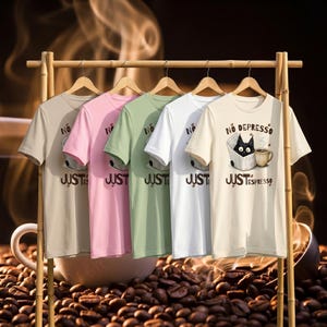 May include: A bamboo clothing rack displays five short-sleeved t-shirts in beige, pink, green, white, and cream. Each shirt has the text "NO" above "JUST" in a bold font. The cream shirt features a black cat in a box with the text "NO DEPRESSO JUST ESPRESSO".