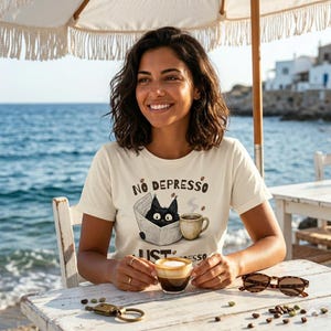 May include: Cream-colored t-shirt with the words "NO DEPRESSO" and "JUST ESPRESSO" printed on it. The graphic features a black cat reading a newspaper and a coffee cup. The person is sitting at a table with a cup of coffee and sunglasses.