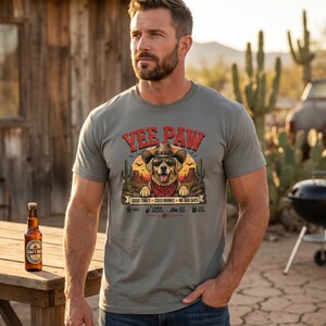 K&ouml;nnte beinhalten: Graues T-Shirt mit einem Aufdruck eines Hundes mit Cowboyhut und Halstuch, mit dem Text "Yee Paw". Das Shirt hat auch den Text "Good Times + Cold Drinks = No Bad Days". Eine Bierflasche steht auf einem Holztisch.