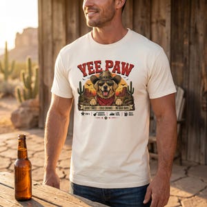 K&ouml;nnte beinhalten: Cremefarbenes T-Shirt mit einem Aufdruck eines Hundes mit Cowboyhut und Halstuch, mit dem Text "Yee Paw". Das Design beinhaltet Kakteen und den Satz "Good Times, Cold Drinks, No Bad Days."