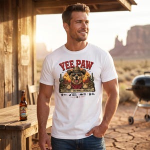 K&ouml;nnte beinhalten: Wei&szlig;es T-Shirt mit einem Aufdruck eines Hundes mit Cowboyhut und Halstuch, mit dem Text "Yee Paw". Das Design umfasst Kakteen und die S&auml;tze "Good Times, Cold Drinks, No Bad Days". Eine Bierflasche steht auf einem Holztisch.