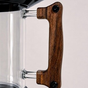 May include: Close-up of a coffee maker with a clear glass carafe and a dark wooden handle. The handle is attached with black screws. The top of the coffee maker is black.