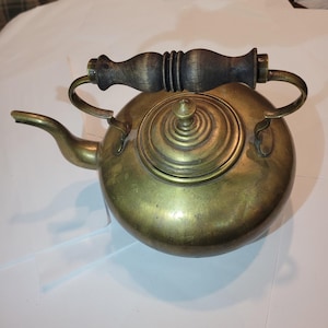 May include: A vintage brass kettle with a round body, a spout, and a wooden handle. The lid is also brass and has a small knob. The handle is dark brown and curved. The kettle is likely used for boiling water.