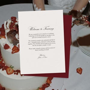 May include: A wedding invitation with the text "Welcome to Tuscany" in elegant script. The invitation is white with a burgundy backing, and the invitation is placed on a cake decorated with strawberries and raspberries.