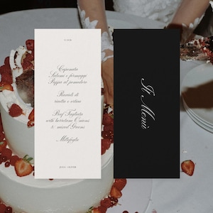Op de afbeelding: Elegante trouwmenukaarten. Eén kaart is wit met een menu in cursief, met gerechten als "Ravioli di ricotta e ortica." De andere kaart is zwart met "Il Menù" in witte cursief. De kaarten liggen bij een witte taart.