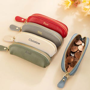 May include: A collection of personalized leather coin purses in red, gray, white, and green, each with a gold zipper and a name or initials. One blue purse is open, filled with pennies. The purses are oval-shaped and have a small leather pull tab.