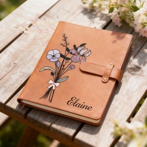 May include: A tan leather journal with a floral design featuring purple and lavender flowers, a pink bow, and the name "Elaine" in script. The journal has a brown leather strap closure.
