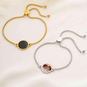 May include: Two adjustable slider bracelets with box chains. One bracelet is gold-toned with a solid black circular center charm. The other bracelet is silver-toned and features a circular center charm displaying a photograph of two people.