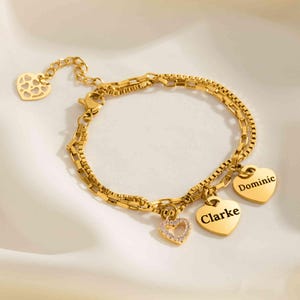 May include: Gold-colored bracelet with heart-shaped charms. One charm has a heart-shaped cutout design, another is encrusted with small crystals, and two others are engraved with the names "Clarke" and "Dominic". The bracelet is on a white surface.