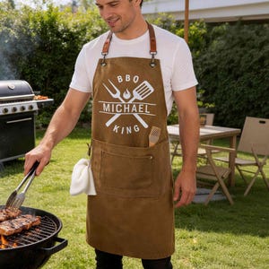 May include: A brown canvas apron with leather straps and a white graphic print featuring a fork, spatula, flame icon, and the text BBQ MICHAEL KING. The apron includes a large front pocket and a white cloth hanging from a side loop.