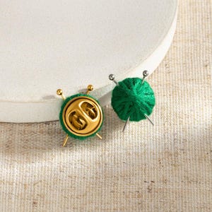 May include: Two green yarn ball pins with silver knitting needles. One pin shows the front with textured green yarn, while the other shows the back with a gold-toned metal clutch backing and two crossed silver needles extending from the sides.