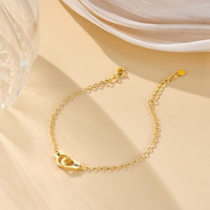 May include: A gold-toned chain bracelet featuring a central charm shaped like a pair of handcuffs. The handcuffs are engraved with the letters K and B. The bracelet includes a delicate link chain and a small circular clasp at the end.
